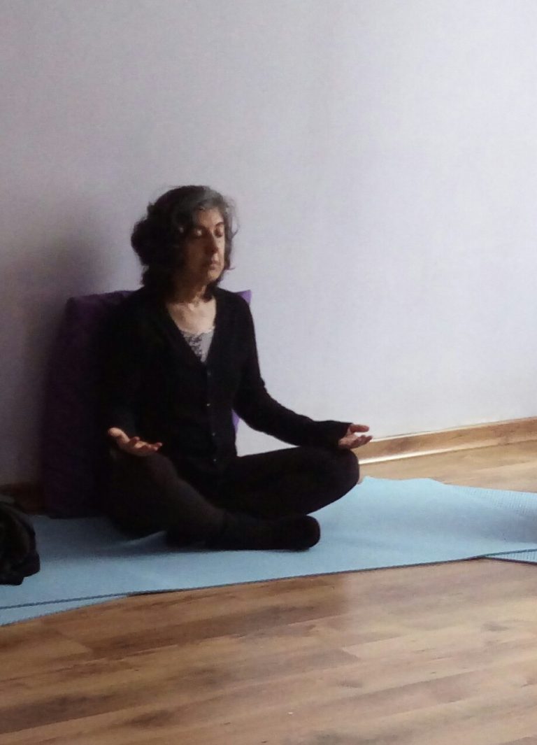 aluna em posição de pernas de índio sentada no tapete azul de Yoga e mãos repousadas sobre os joelhos de palmas para cima e olhos fechados