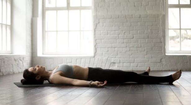 mulher deitada no chão de olhos fechadas com calça preta e top cinza realizando meditação
