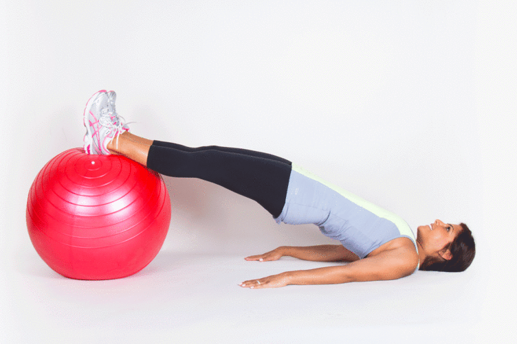 moça jovem com calça preta e blua azul claro puxando bola vermelha com os pés e quadris elevados