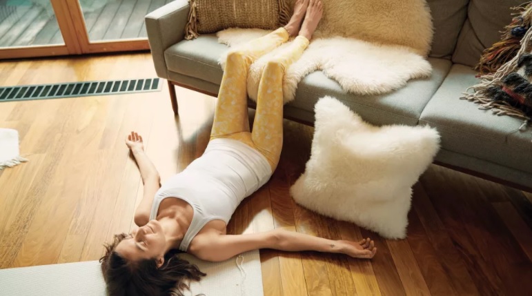 mulher com blusa branca e calça amarela deitada no chão com os braços abertos e as pernas apoiadas no sofá cinza com manta de pelúcia branca e almofada de pelúcia branca no chão apoiada em pé no sofá