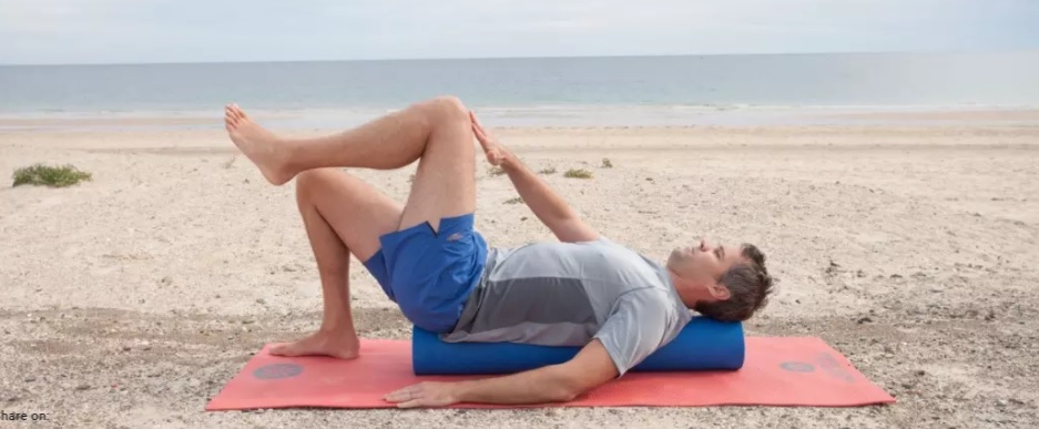 homem de meia idade com shorts azul claro e camiseta cinza deitado em rolinho na vertical em tapete rosa tocando a mão direita em perna esquerda com joelhos flexionados, ambiente de praia
