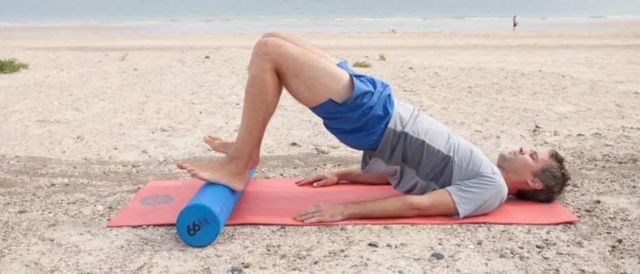 homem de meia idade com shorts azul claro e camiseta cinza deitado em rolinho na vertical em tapete rosa, pés apoiados no rolinho na horizontal, joelhos flexionados, quadril elevado, ambiente de praia