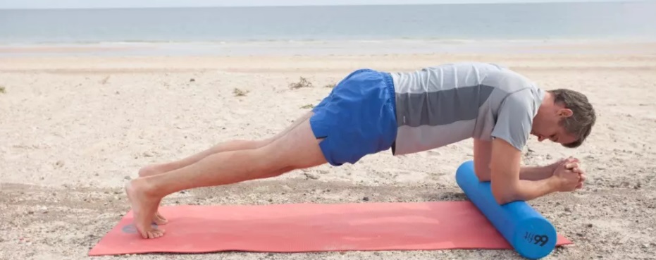 homem de meia idade com shorts azul claro e camiseta cinza deitado em rolinho na vertical em tapete rosa, com antebraços apoiados no rolinho na horizontal, pernas estendidas com pés em meia ponta apoiados no solo, em prancha baixa, ambiente de praia