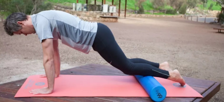 Homem de meia idade com calça preta e camiseta cinza com canelas apoiada no rolinho em posição de prancha alta