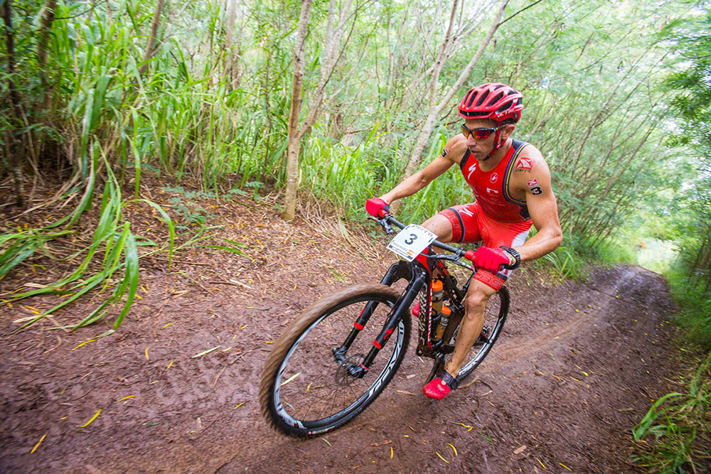 ciclista com roupa laranja em moutain bike em estrada de terra com mato ao fundo