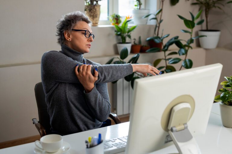 Pessoa sentada em frente ao computador no home office fazendo um alongamento com os braços aprendido no pilates, com expressão de alívio, demonstrando cuidado com a coluna.